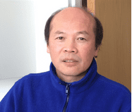 A middle-aged man with short black hair and a receding hairline is wearing a blue zip-up jacket, sitting indoors with a neutral expression.