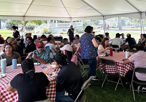 A group of people sits at round tables covered with red and white checkered tablecloths under a large outdoor tent, eating and talking at a picnic event. Trees and a gazebo are visible in the background.