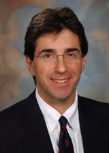 A man with short dark hair and glasses, wearing a dark suit, white shirt, and patterned tie, smiling at the camera against a blurred background.
