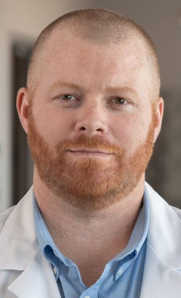 A man with a red beard and short hair, wearing a white lab coat over a blue collared shirt, looks directly at the camera with a neutral expression.