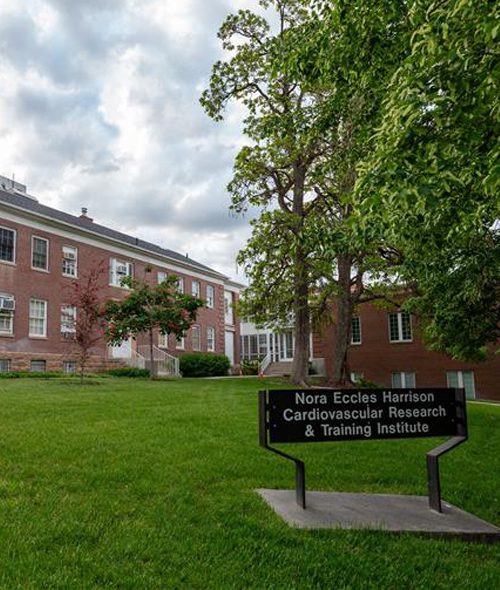 A grassy lawn with trees in front of a brick building. A black sign in the foreground reads “Nora Eccles Harrison Cardiovascular Research & Training Institute.” The sky is partly cloudy.