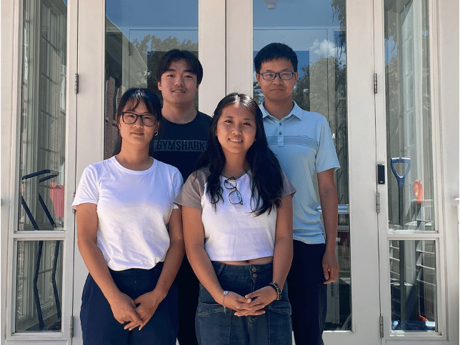 Four young adults stand in two rows, posing for a photo in front of large glass doors on a sunny day. Two women are in front, and two men are behind them.