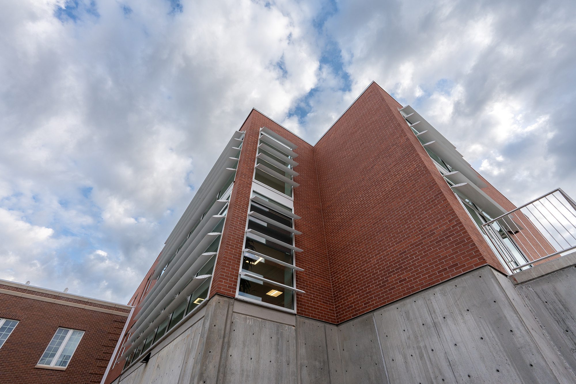 A modern building with red brick walls, concrete base, and horizontal white metal slats is viewed from below against a partly cloudy sky.