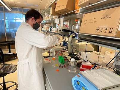 A person in a white lab coat and mask works at a laboratory bench with scientific equipment, pipettes, and containers, surrounded by boxes and lab supplies.