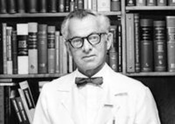 A man wearing glasses, a white coat, and a bow tie stands in front of bookshelves filled with books.